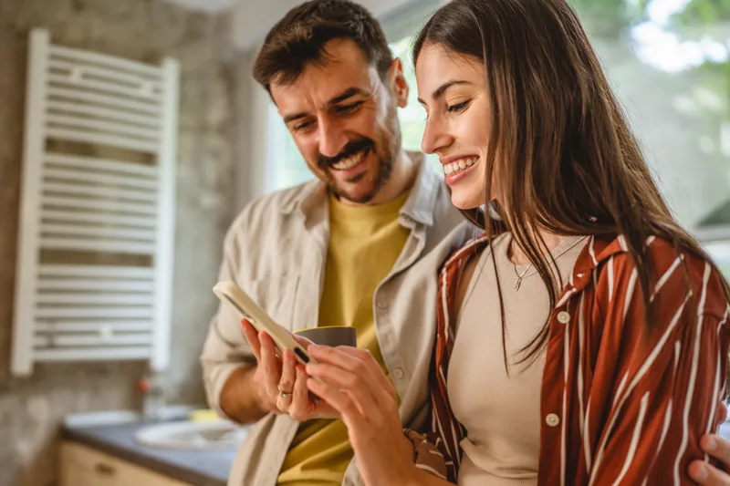 A smiling couple in their kitchen look at a phone together, paying a bill from the app