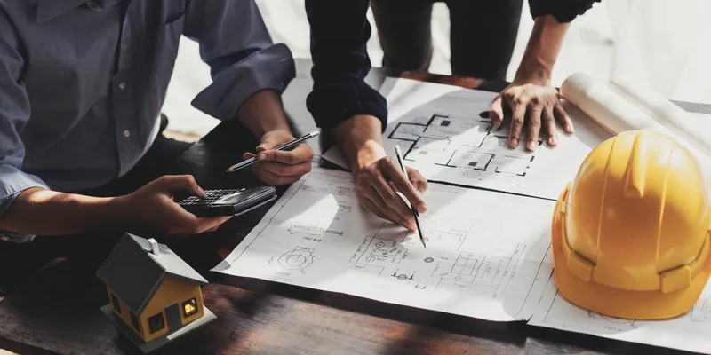 Two people reviewing construction drawings with a pencil and a phone next to a yellow hard hat