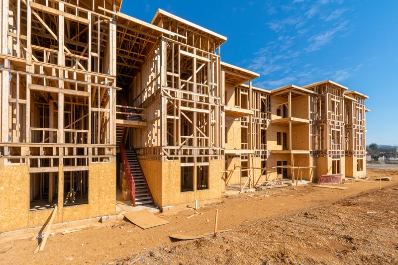 Wood-framed apartment buildings under construction on a dirt lot under a blue sky