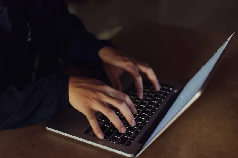 Hands typing on a laptop keyboard in a dark room, lit only by the screen